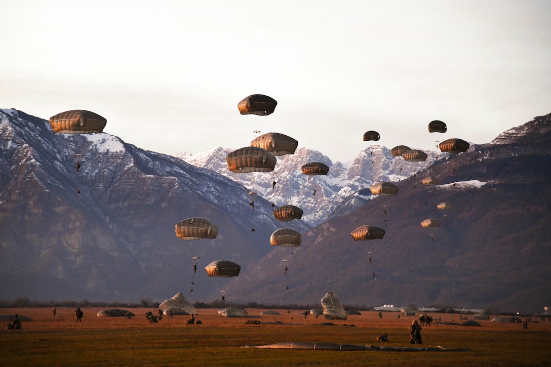 Soldiers freefall wearing parachutes near mountains.