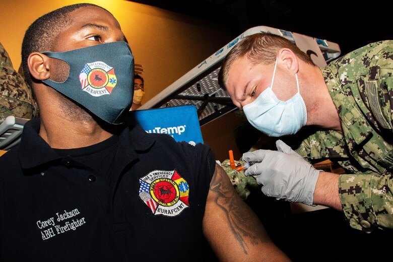 A Navy petty officer wearing a face mask and gloves gives a COVID-19 vaccine to a Navy seaman.