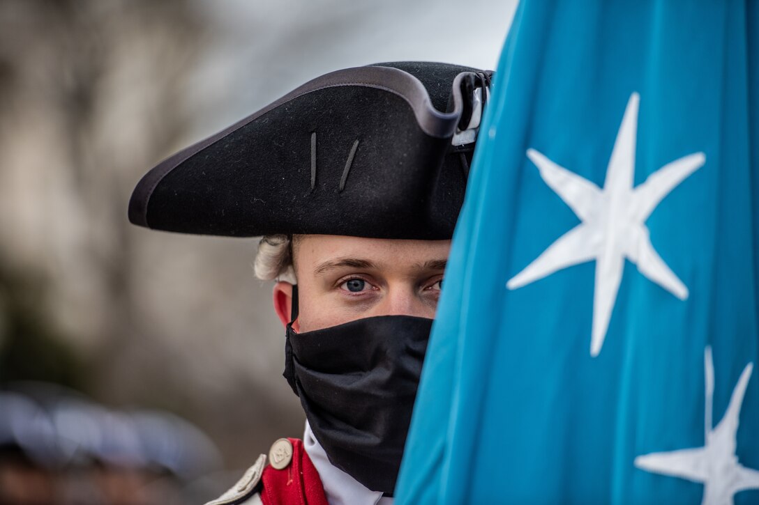 A soldier dressed in a colonial military uniform holds a flag.