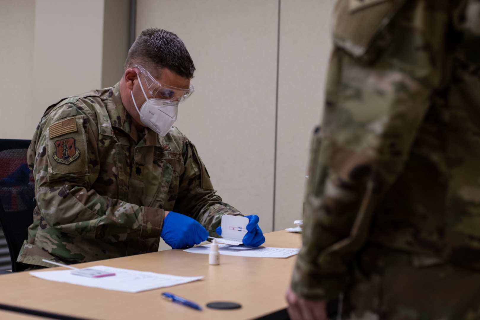A military member in uniform and personal protective equipment works with equipment on a table.