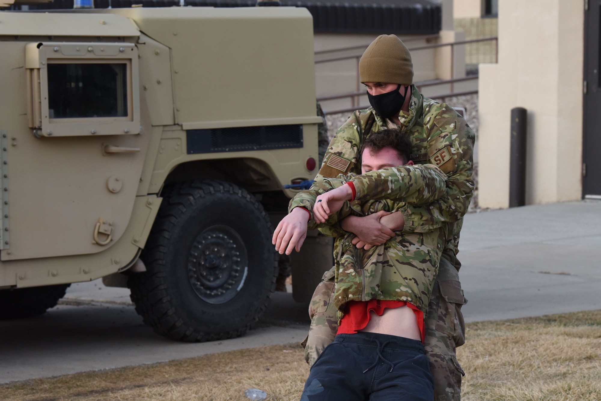 An Airman grips an injured Airman around the chest and drags him backwards behind a Humvee for cover during a simulated training exercise.