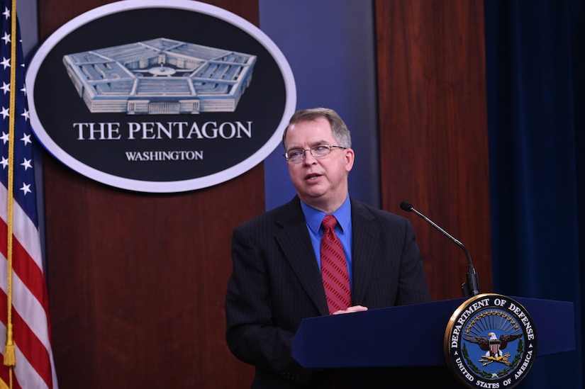 A man stands at a podium in front of a microphone with a large flag on a stand to his right; an oval sign on the wall behind him bears an image of a five-sided building and the words "The Pentagon".