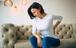 A woman sitting on a couch holding her lower back in pain.