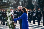 A woman and an older man put their hands on a large wreath that sits on a stand.