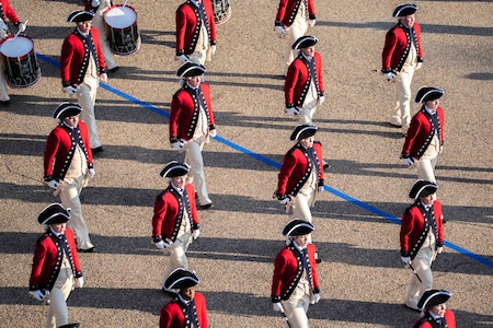 A group of soldiers walk in formation; some hold drums.