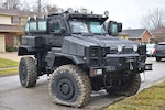 An MRAP with bullet marks and flat front tires is parked on a residential street.