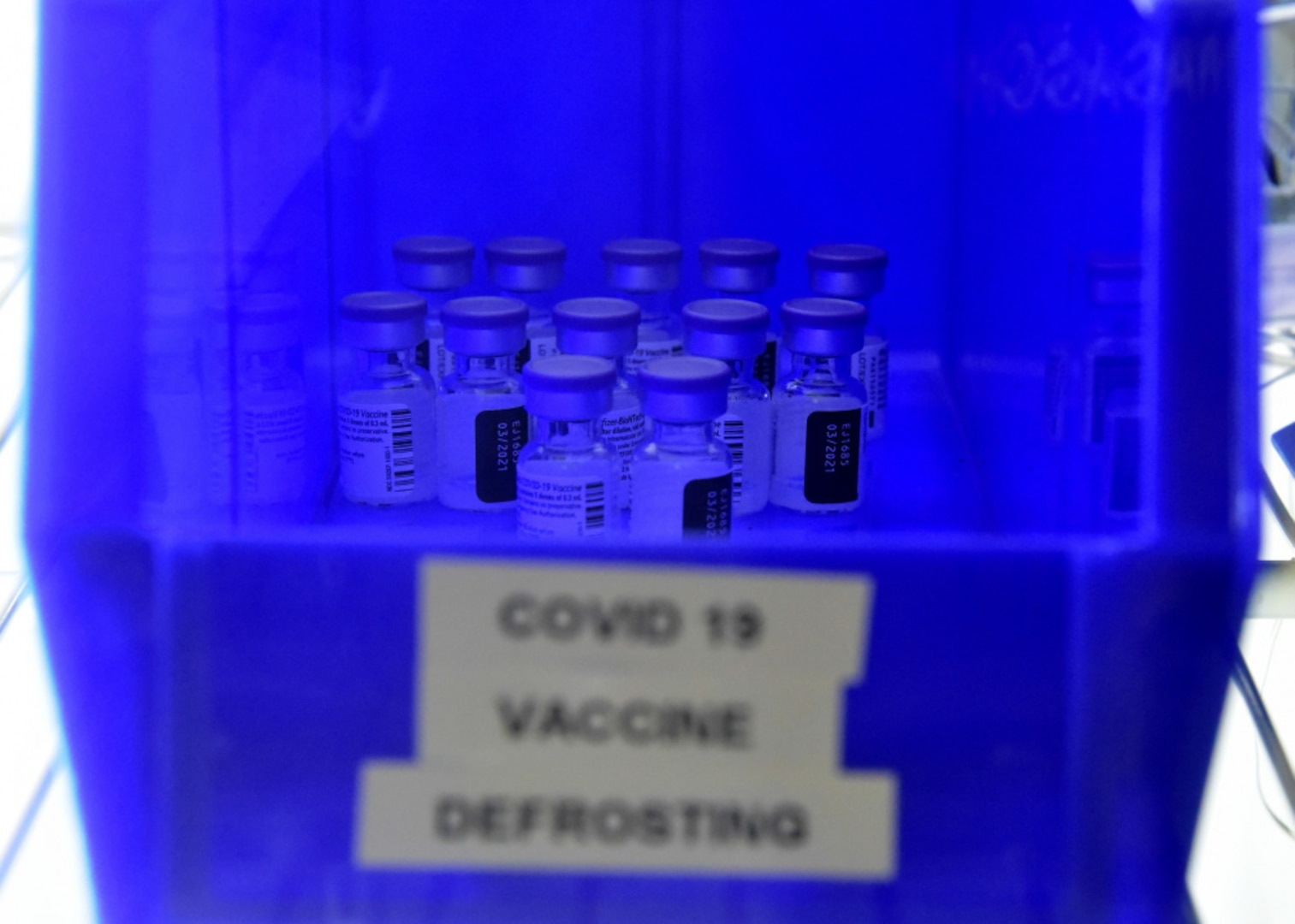 A blue container of vials labeled COVID-19 Vaccine Defrosting sits center frame.