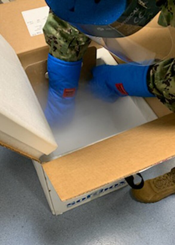 A servicemember in Navy warfare uniform and protective equipment reaches into a box from which fog is emerging from dry ice.