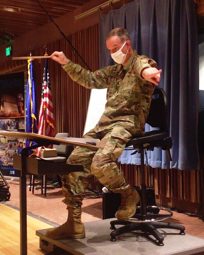 Lt Col Michael Willen conducts a socially distanced Academy Band  rehearsal for our 2020 Holly and Ivy program.  He is dressed in dark green OCP uniform, wearing a white facial mask in front of a blue curtain backdrop as well as the American and Air Force flags. 
 This full program is available for all to watch for free at https://www.youtube.com/channel/UCjCwDVD-ocZPdws2tvIvYog.