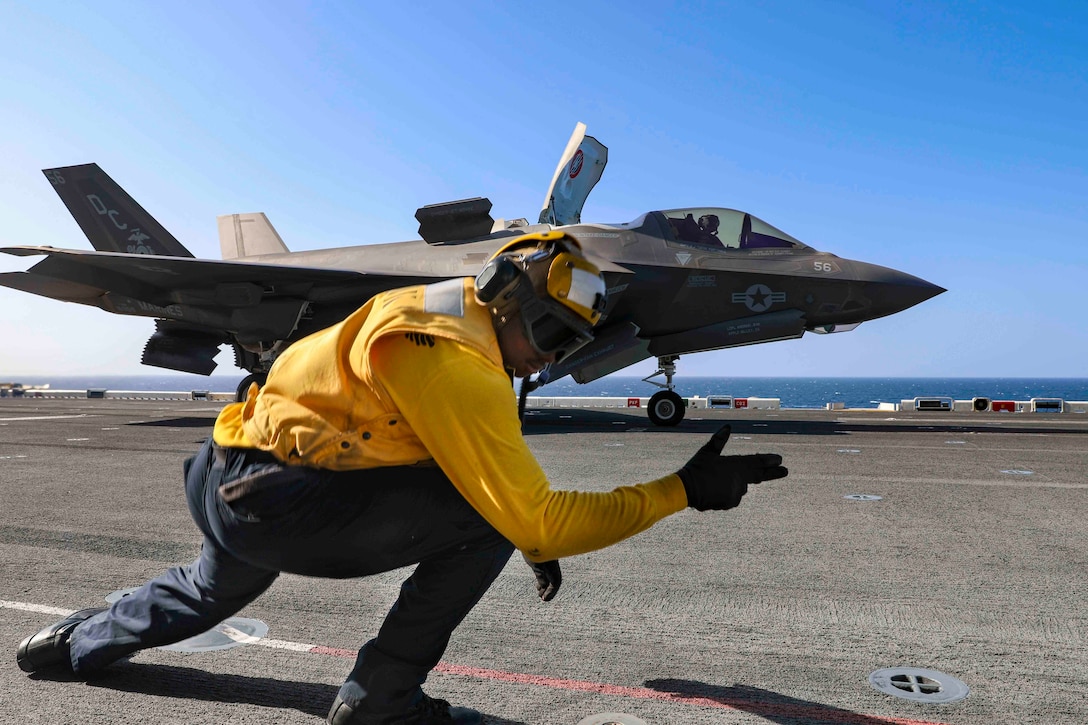 A sailor kneels on the ground signaling toward an aircraft.