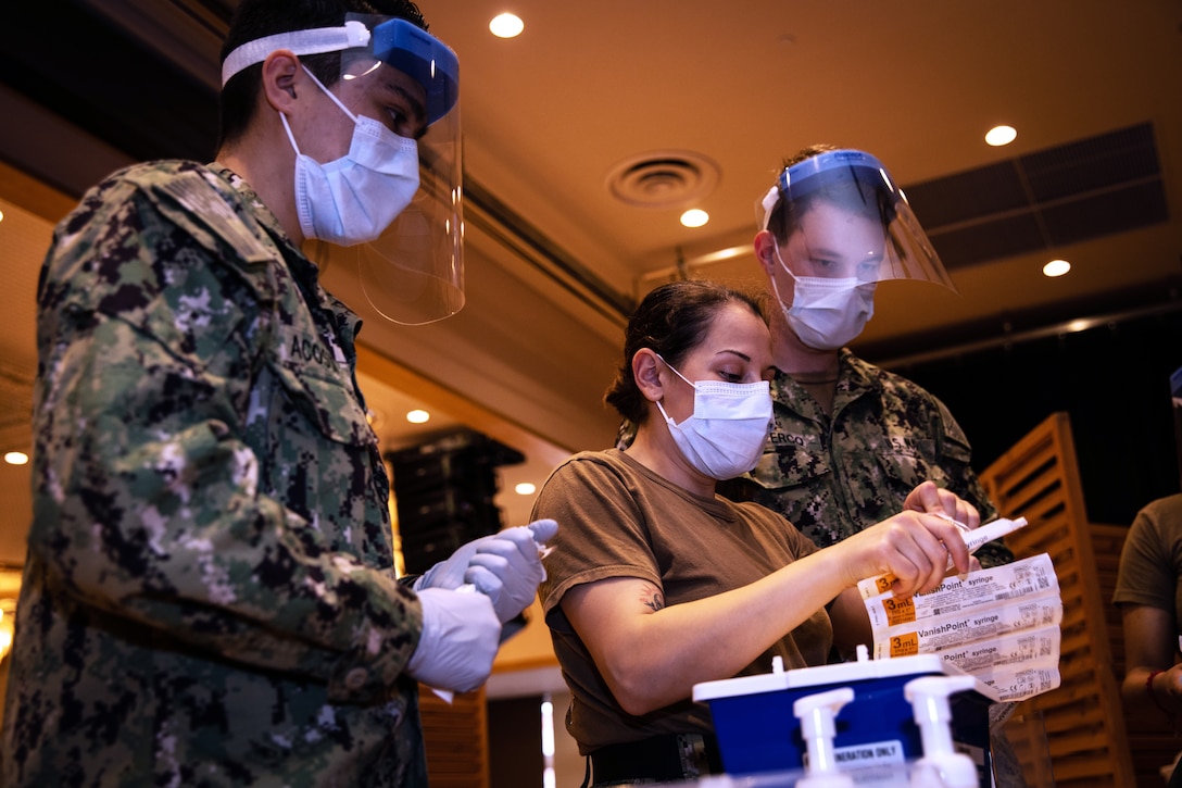 Several service members wearing face masks and gloves prepare to administer COVID-19 vaccines.