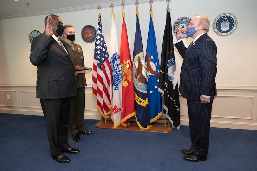 Defense Secretary Lloyd J. Austin III stands next to a man holding up his right hand facing another man.
