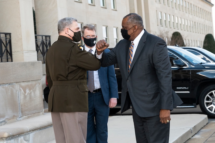 Two men wearing protective face masks bump elbows in front of the Pentagon entrance.