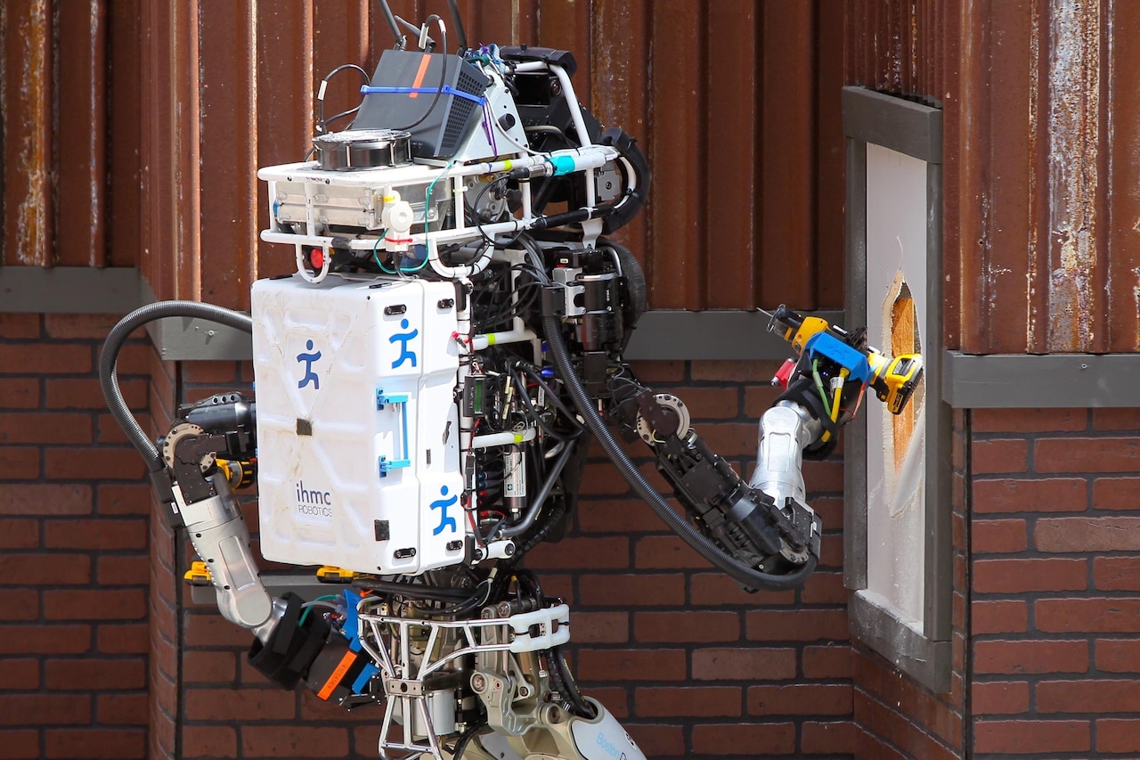 A person-size robot holds a piece of equipment used to cut through drywall on the side of a building.