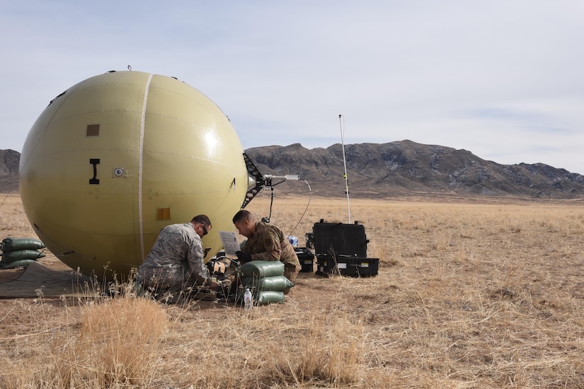 Two men dressed in military uniforms kneel on the ground to work on satellite communications; mountains are in the background.