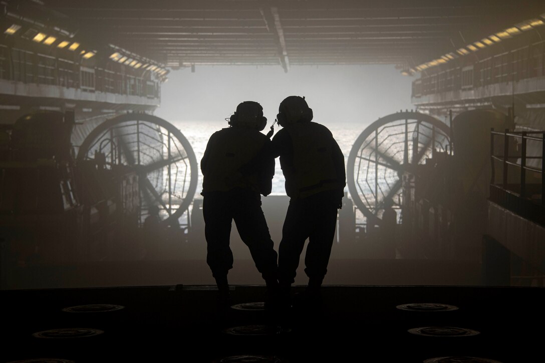 Two sailors shown in silhouette face the ocean while standing on a ship’s deck.