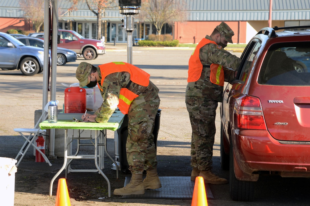A guardsman wearing a face mask reaches into the driver's side window of a vehicle while another guardsman wearing a face mask writes on a clipboard.