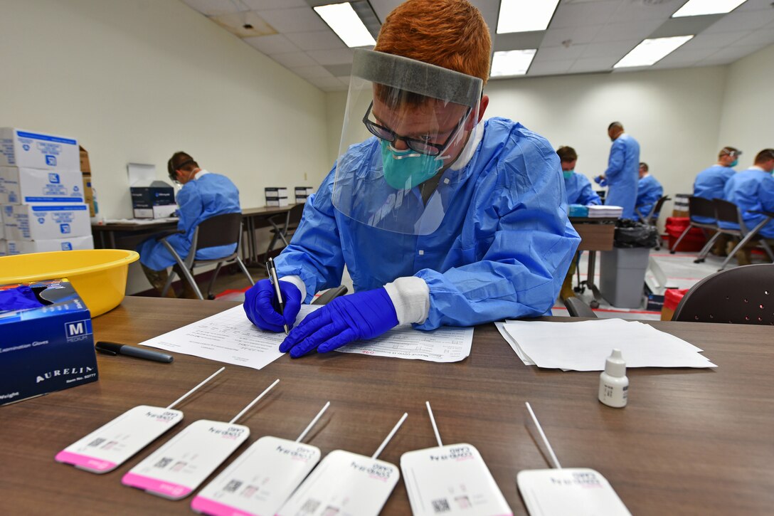 A person wearing personal protective equipment sits at a table and writes on a piece of paper.
