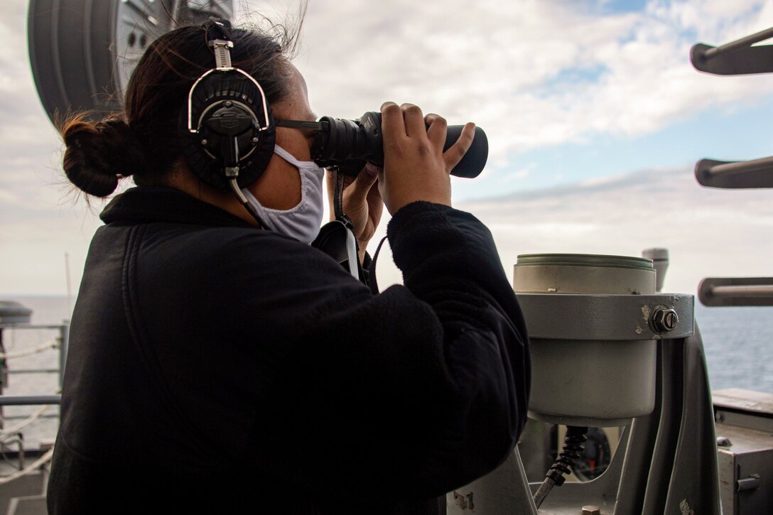 A sailor uses binoculars while standing on a ship at sea.