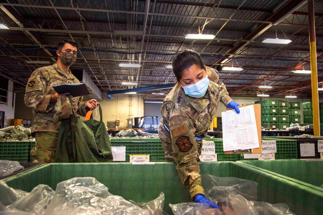 An airman reaches into a bin for supplies while another service member stands nearby holding a bag.