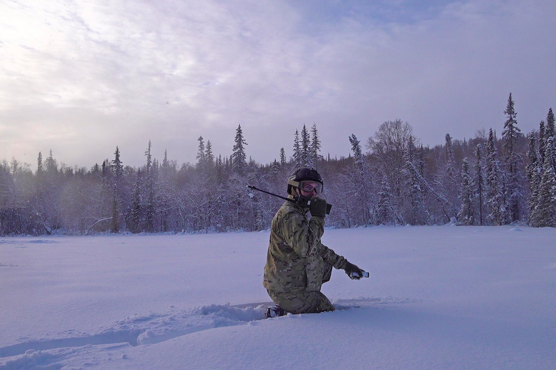 An airman speaks into a radio while kneeling in snow in front of a tree line.