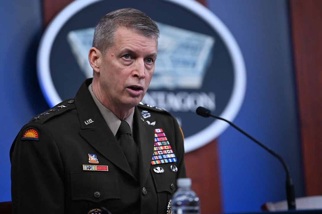 Man in military uniform speaks into a microphone. A sign indicating that he is at the Pentagon is in the background.