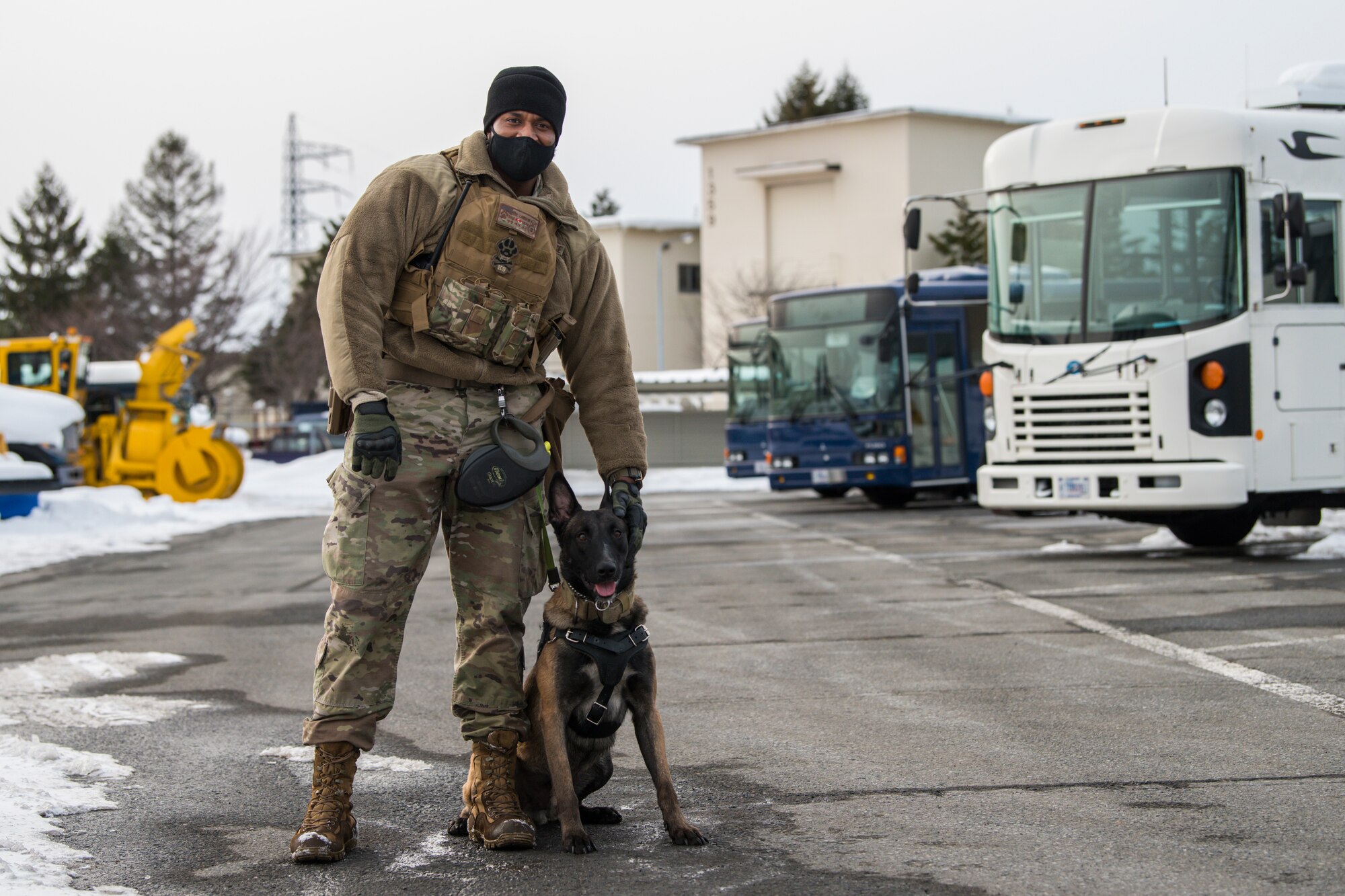 Man in uniform stands in a snowy parking lot next to his sitting dog, while they both look at the camera.