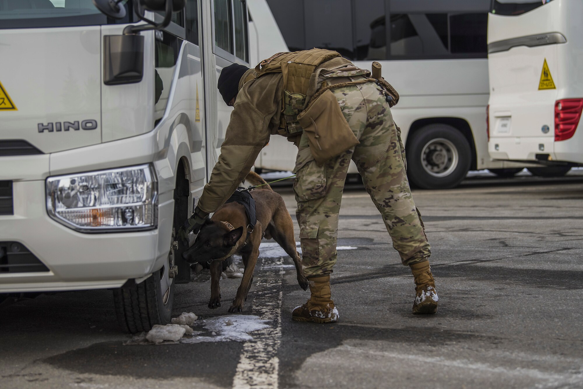 Man in uniform points to a wheel of a vehicle while the dog sniffs for odors next to it.