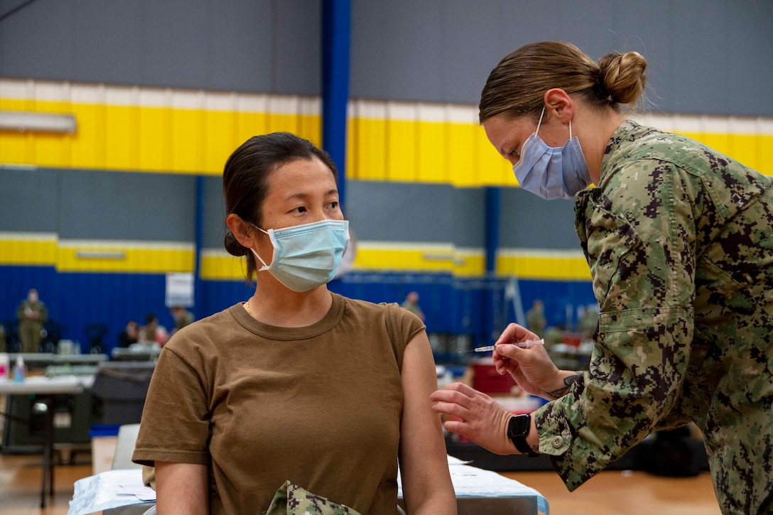 A Navy sailor wearing a face mask administers a  vaccine to another sailor.