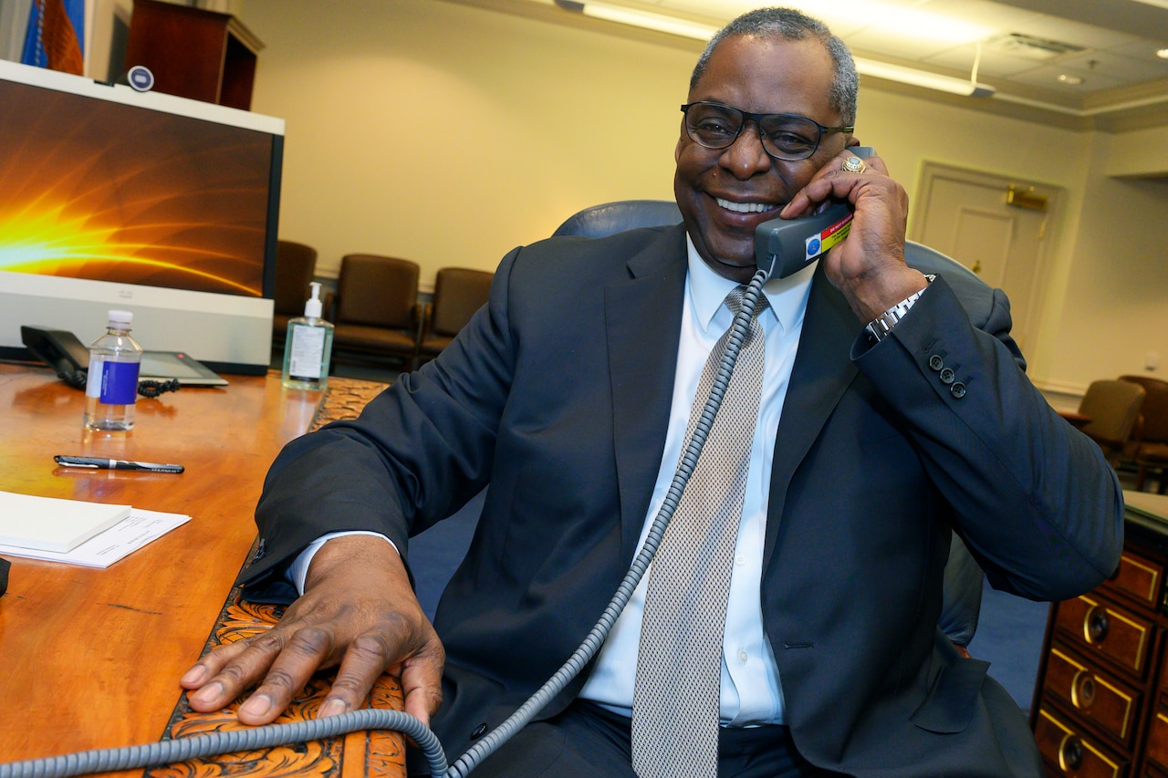 Secretary of Defense Lloyd J. Austin III smiles while talking on a phone at a desk.