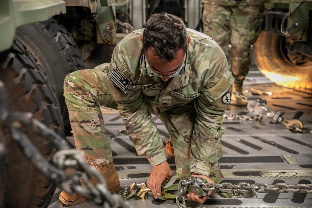 A soldier kneels as he uses a chain to secure a humvee inside a military aircraft.