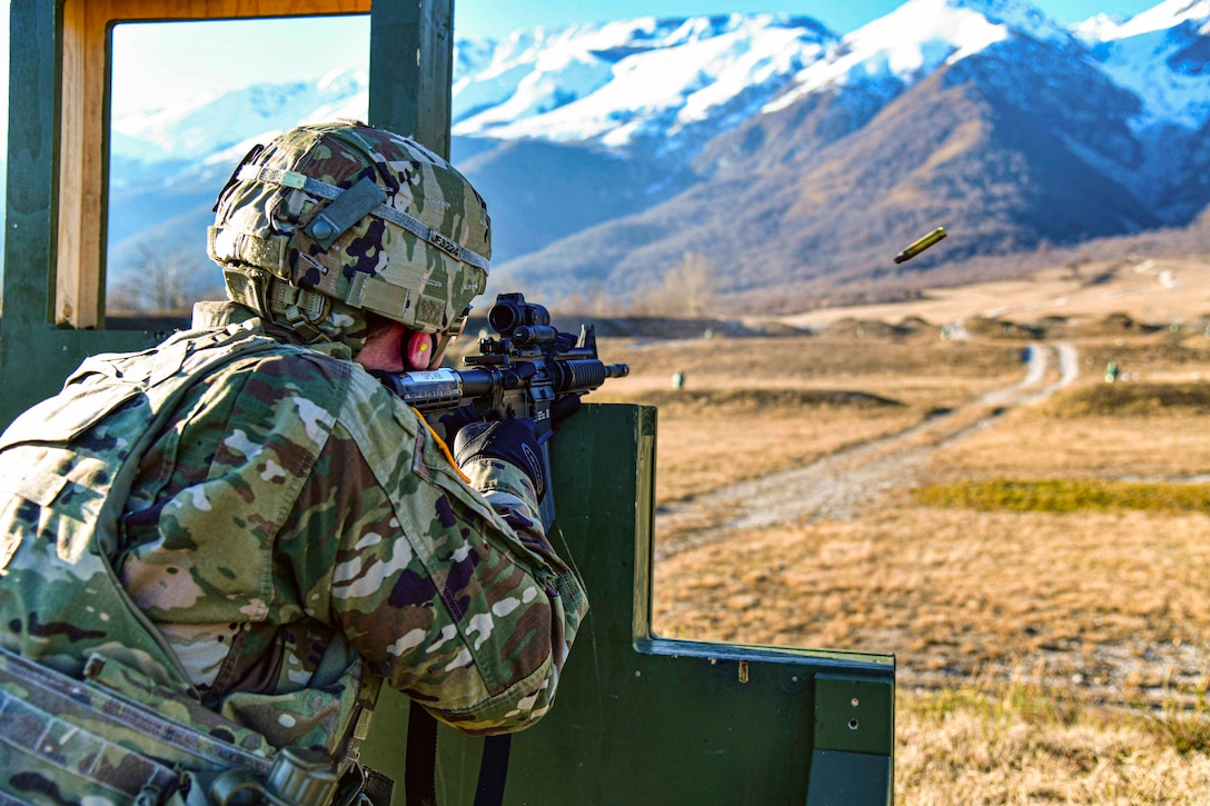A soldier fires a weapon at targets from behind an obstacle; a mountain can be seen in the foreground.