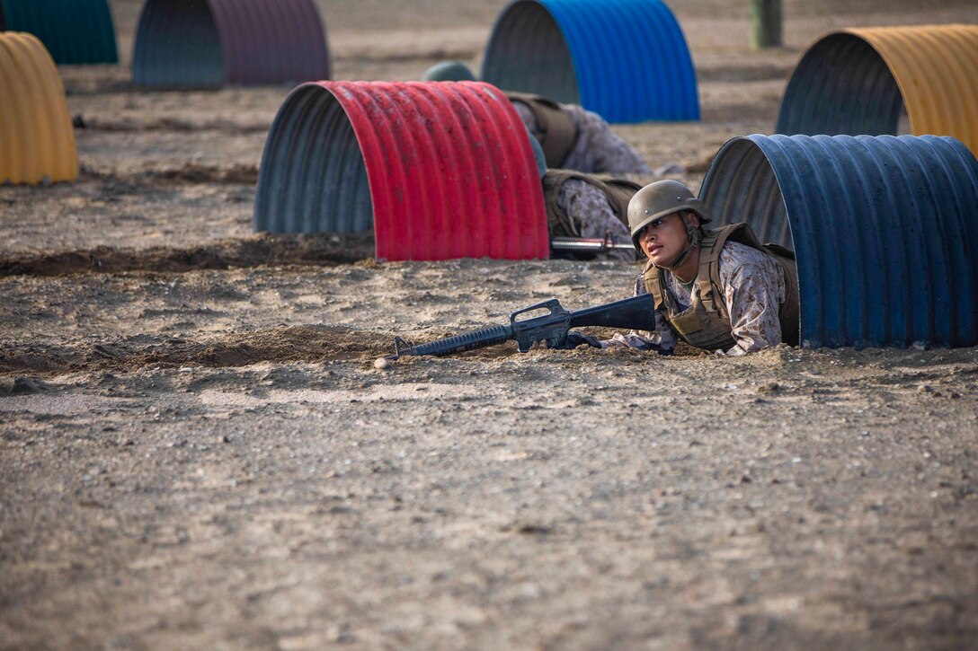 A Marine Corps recruit crawls through a barrel while holding a weapon.