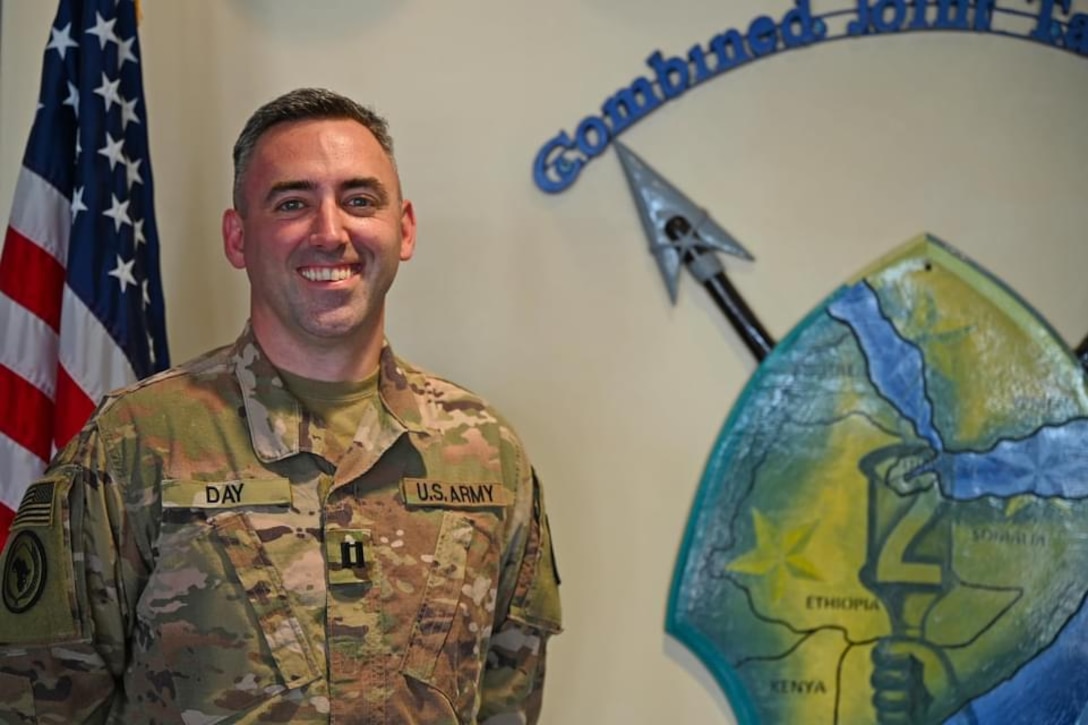 A man in combat uniform smiles beside a unit logo.