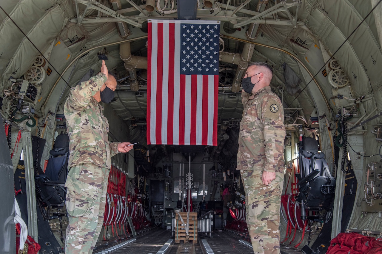 Two men facing each other hold up their right hands inside a C-130 airplane. A U.S. flag hangs in the background.