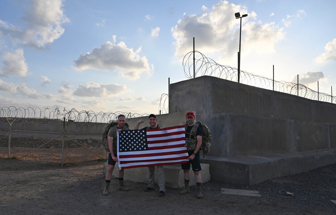 Three men holding a U.S. flag stand near barbed-wire fences.