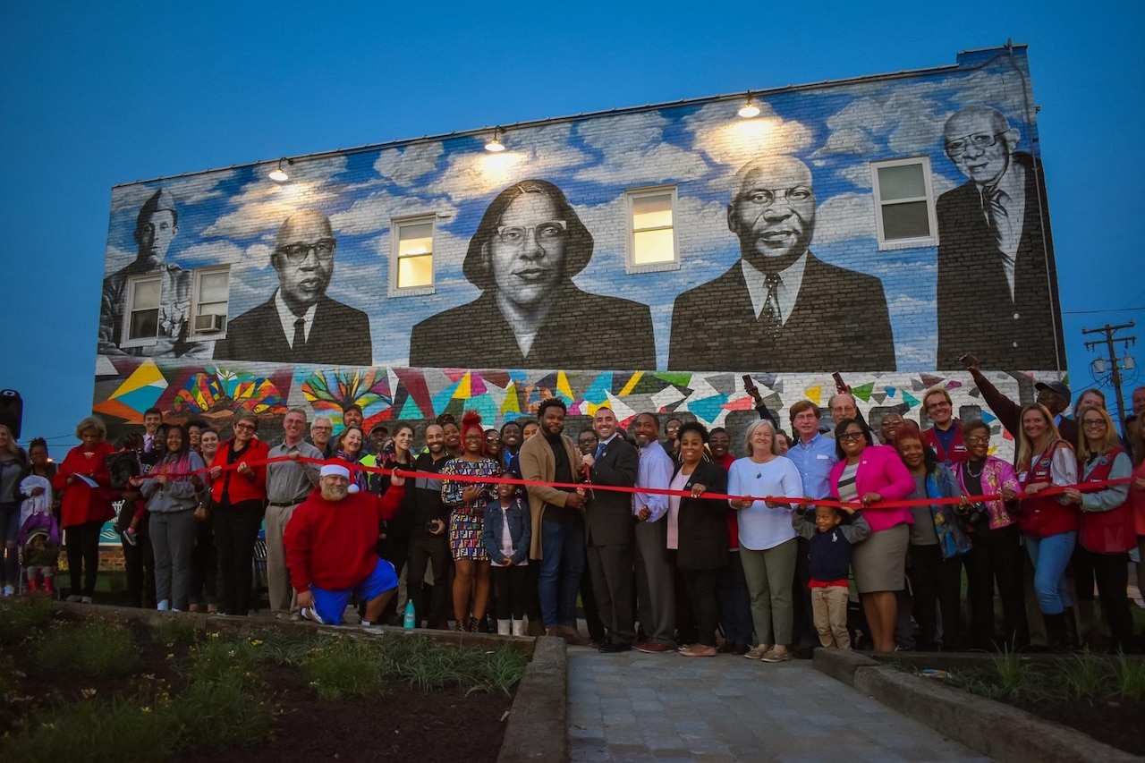 A crowd prepares to cut a ribbon in front of a mural on a building.