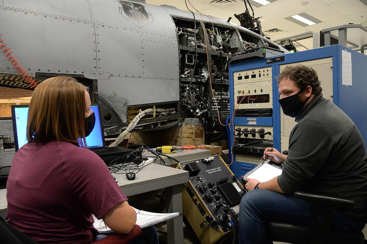 Two people, amid an array of electronics, talk.