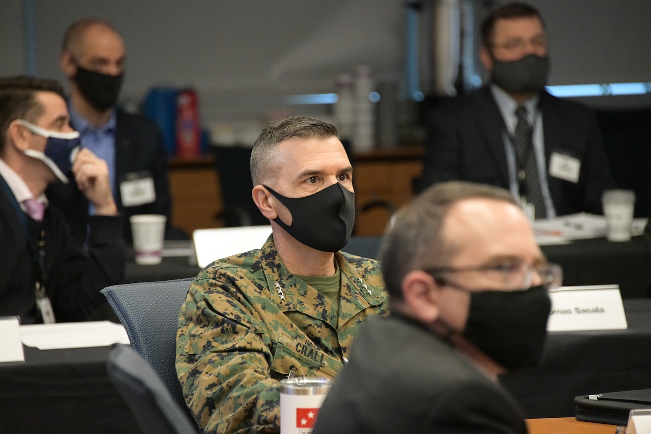 A man in uniform and wearing a mask observes a meeting.