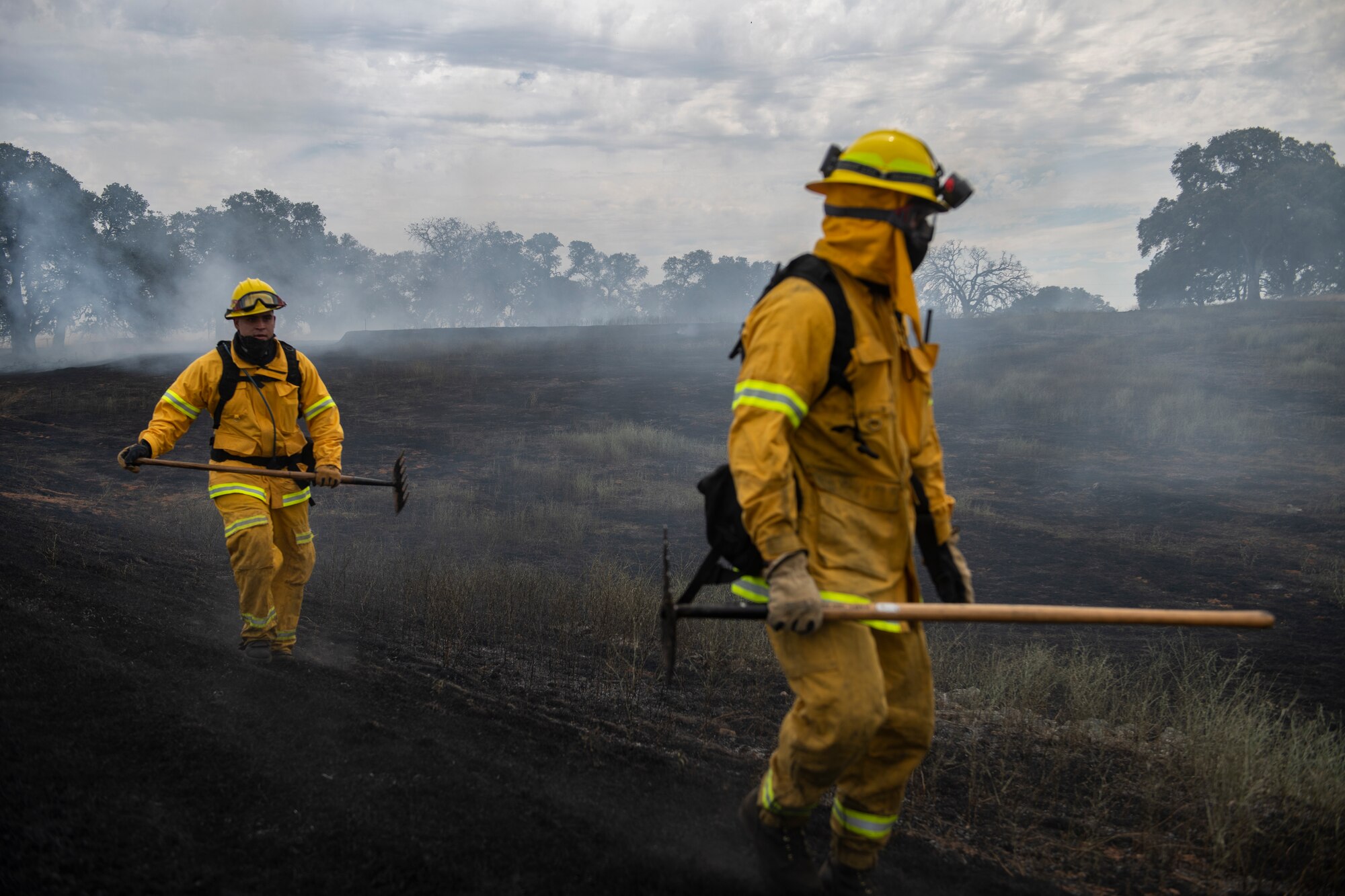 Two firefighters walk through a charred landscape holding axes.