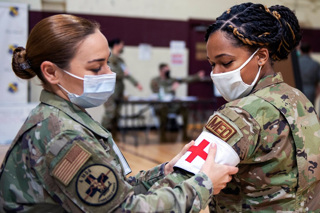 An airman wearing a face mask places a red cross armband around the upper arm of another airman wearing a face mask.
