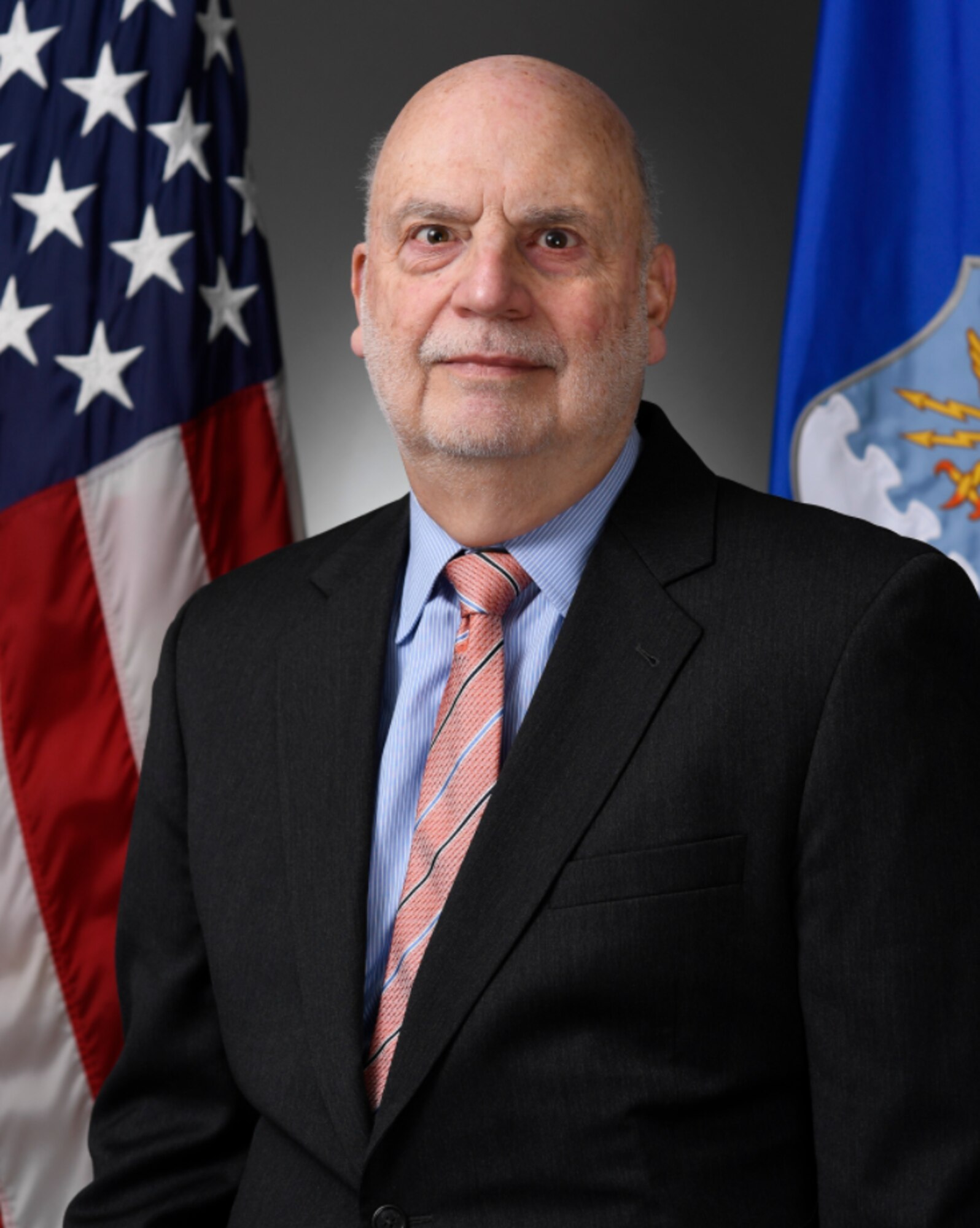 Acting Secretary of the Air Force John Roth poses in front of an Air Force flag on his left, and an American flag on his right. He looks tired.