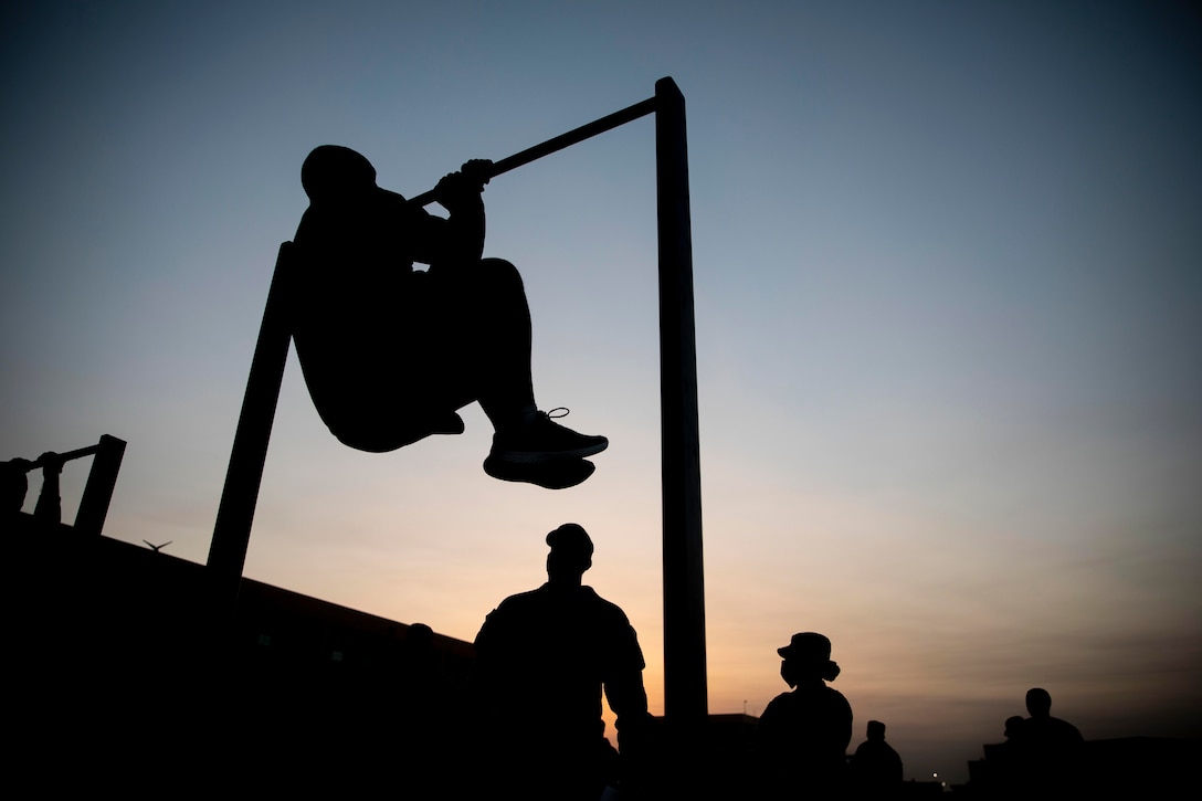 A soldier shown in silhouette does a leg tuck on a bar as others watch.