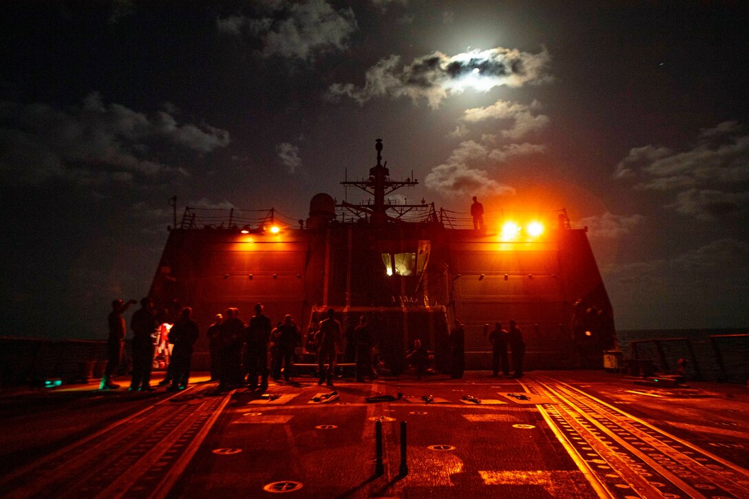 Sailors gather on a ship’s deck at night illuminated by stadium lights.