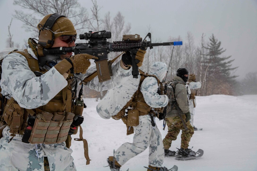 A Marine looks through the scope of a weapon as others move behind in the snow.