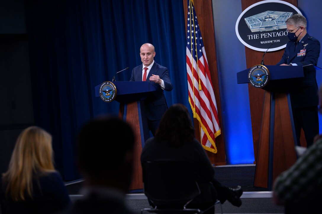 Two men stand behind lecterns and speak to a group of people.