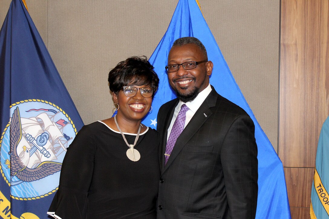 A man and woman stand in front of military flags.