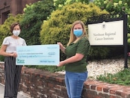 Photo of two women standing outside, holding a large square faux check.  One woman has short dark hair; the other has long blonde hair.  Behind them, there is a sign that reads, "Northeast Regional Cancer Institute."