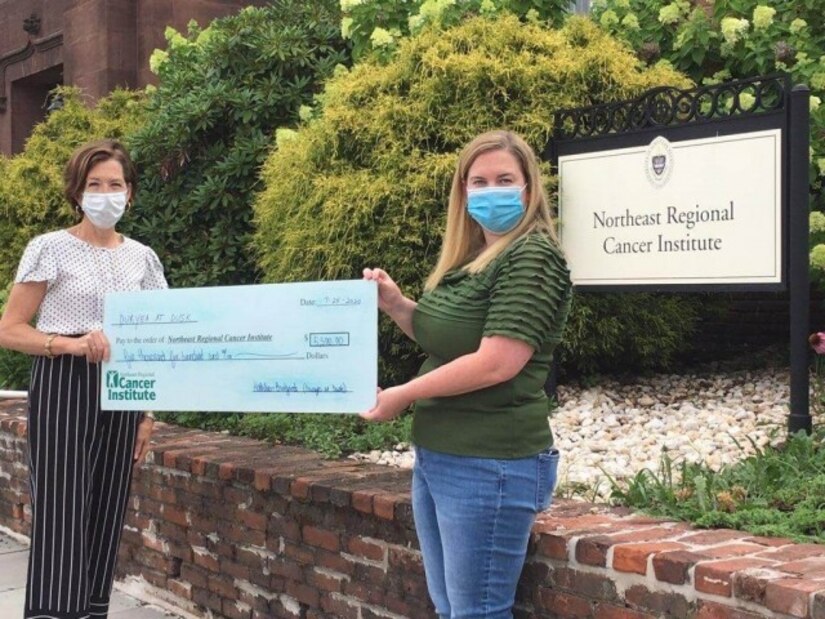 Photo of two women standing outside, holding a large square faux check.  One woman has short dark hair; the other has long blonde hair.  Behind them, there is a sign that reads, "Northeast Regional Cancer Institute."