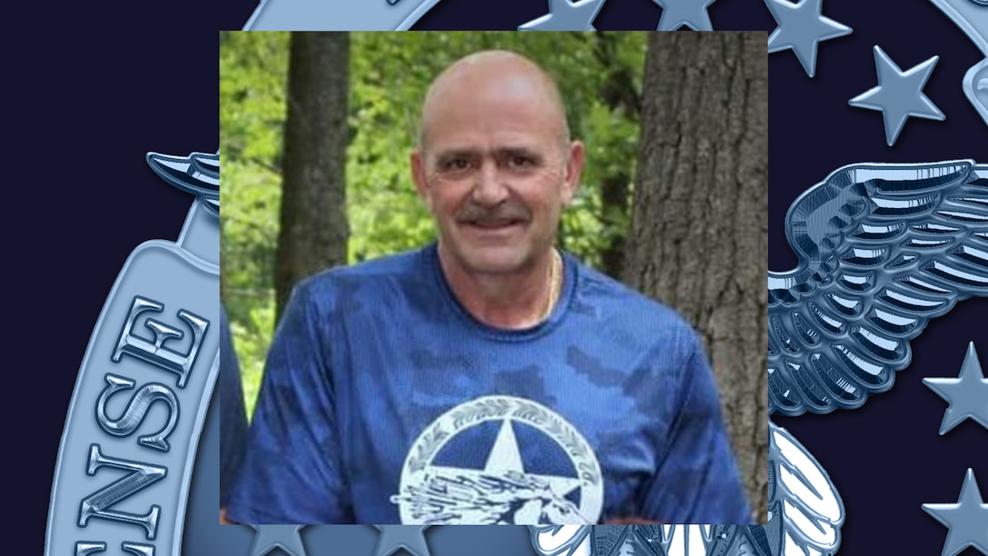 Mark Aicher wearing blue shirt with trees in the background overlaid on a navy blue backdrop